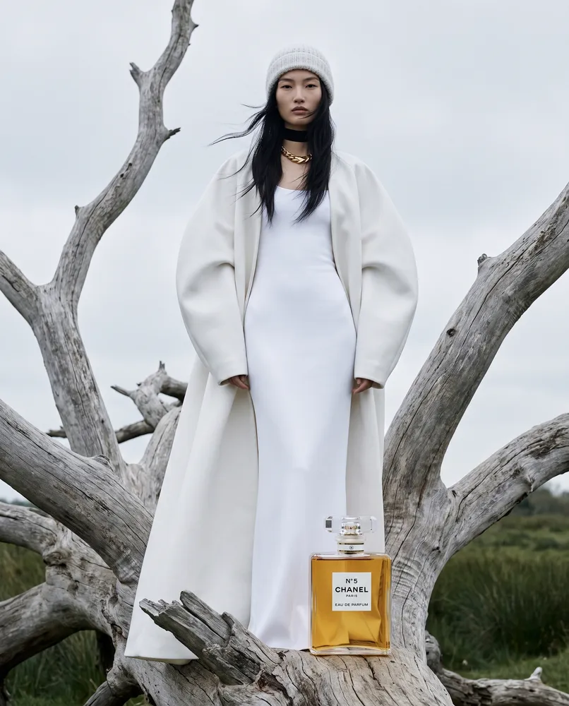 Woman in white coat with perfume by driftwood — AI · The o.KI Agency